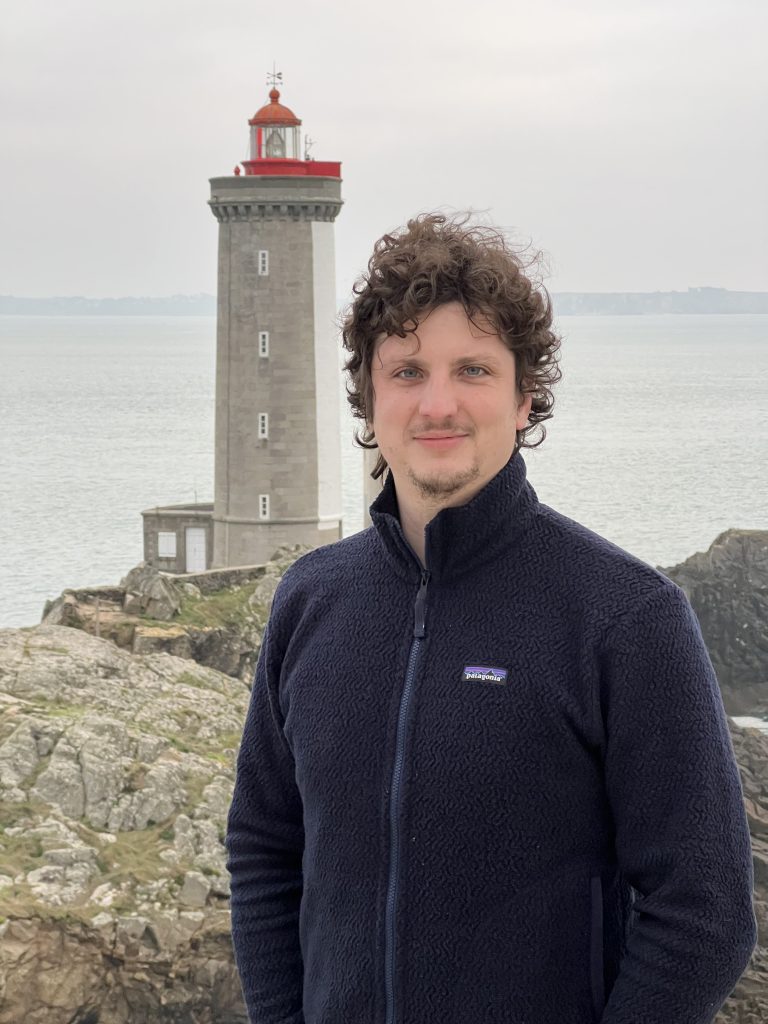 Alessio Andreani con un pile scuro, davanti a un faro sulla costa della Bretagna.